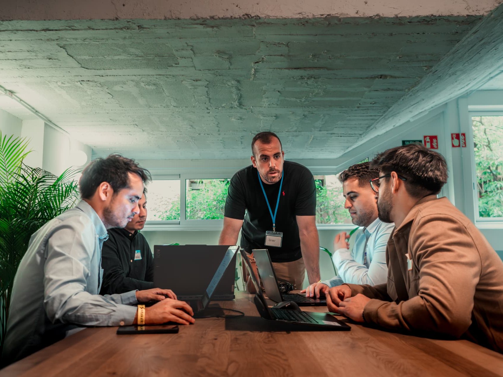 A person wearing a name badge standing and guiding four people working on laptops at a long table