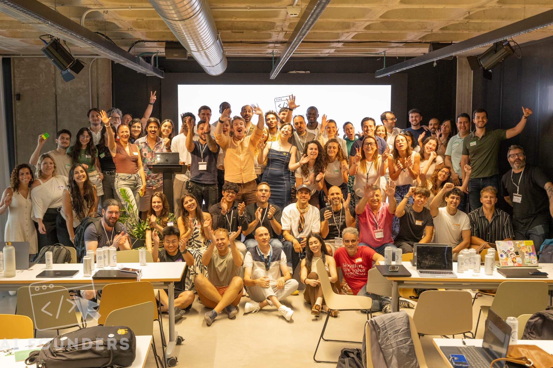 Around fifty attendees and organizers posing together for a group photo at the venue, laptops open on the desks in front